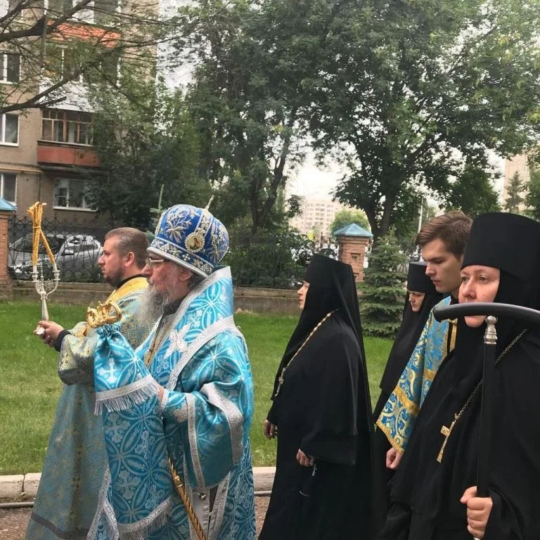 Митрополит в голубом облачении с жезлом идёт по территории храмового комплекса в сопровождении духовенства