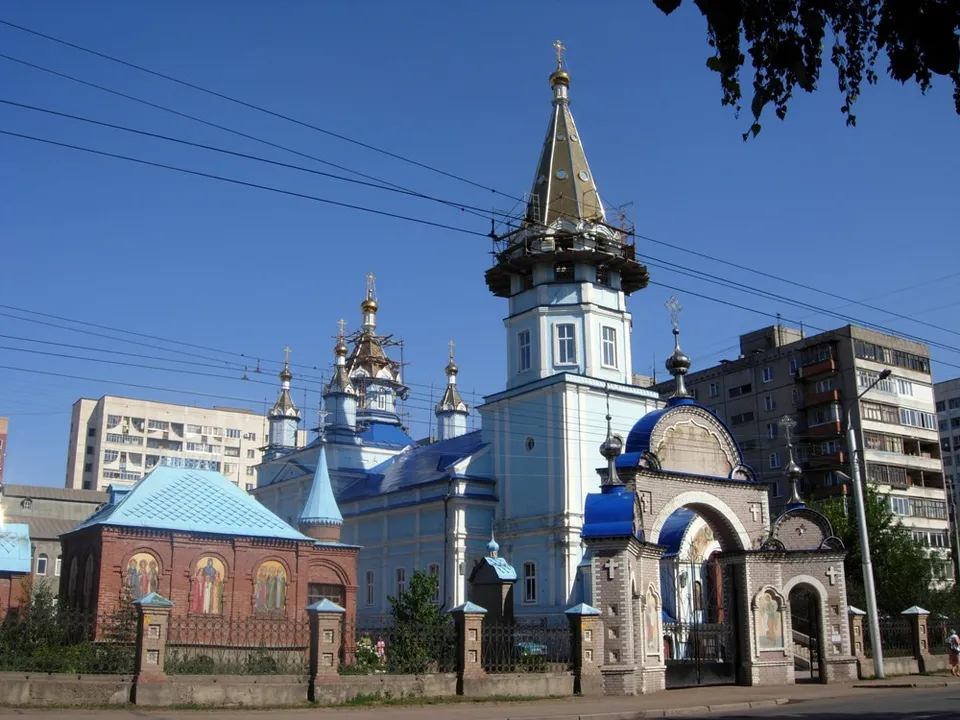 Кирпичный храм с голубыми куполами и шатровой колокольней среди зелени